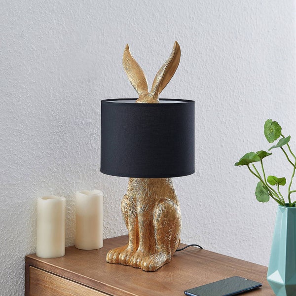 Tischleuchte in Form eines goldenen Hasen mit schwarzem Lampenschirm auf einem Holztisch