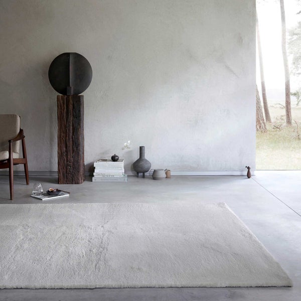 Großer, heller Teppich in einem minimalistischen Raum mit Betonboden, strukturierter Wand und dekorativer Holzskulptur.