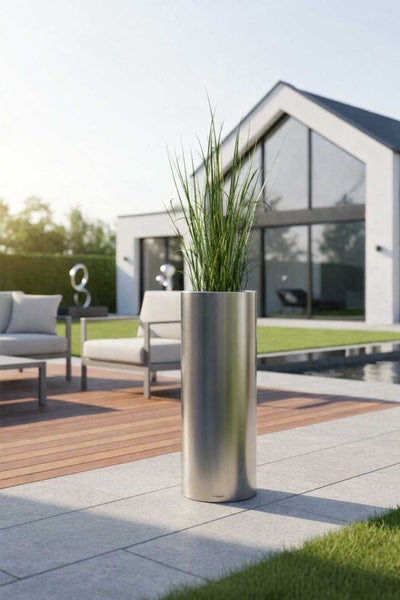 Hoher zylindrischer Metall-Pflanzkübel mit Ziergras auf einer modernen, hellen Terrasse mit Holzdeck, Loungemöbeln und Pool vor einem Wohnhaus.