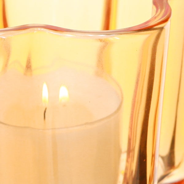 Nahaufnahme einer weißen Kerze mit zwei brennenden Dochten in einem gewellten Windlicht aus bernsteinfarbenem Glas.