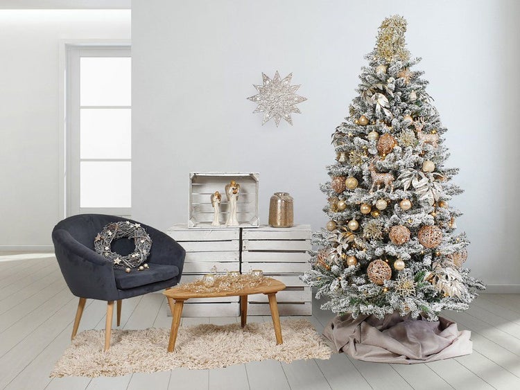 Weihnachtliche Wohnszene mit geschmücktem, beschneitem Weihnachtsbaum, Samtsessel, Holzkisten, Tisch und Deko in festlicher Atmosphäre.