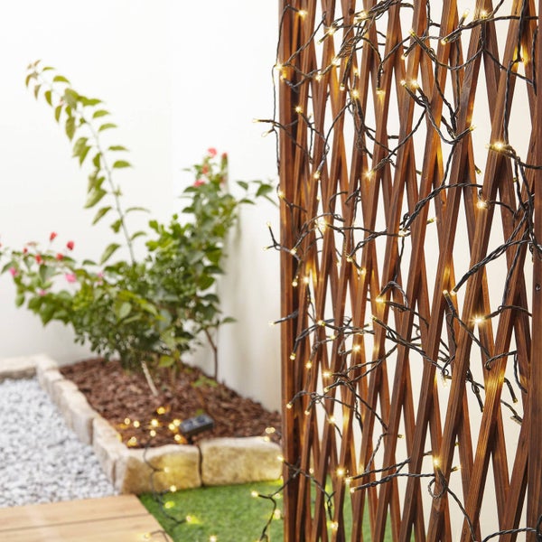 LED-Lichterkette mit schwarzem Kabel an einem hölzernen Rankgitter in einem Garten mit Pflanzen und Steinumrandung.