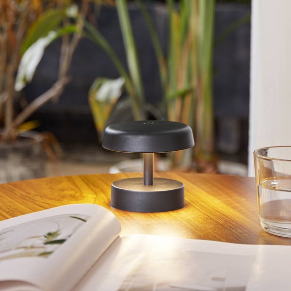 Schwarze kabellose LED-Tischleuchte auf einem Holztisch neben einem Buch und einem Glas Wasser.