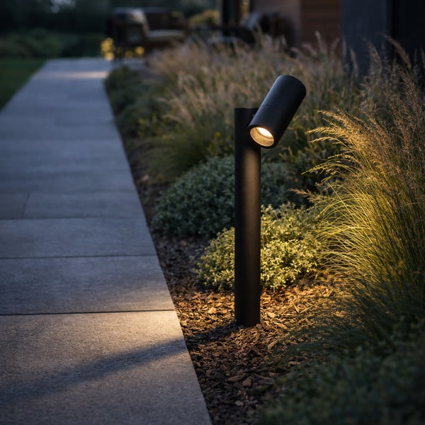 Moderne schwarze Pollerleuchte mit verstellbarem Spot an einem Gartenweg zwischen Gräsern und Mulch.