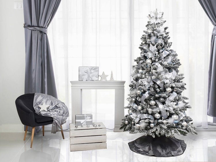 Weihnachtsbaum mit künstlich verschneiten Zweigen, dekoriert mit silbernen Kugeln, Bändern und Blumen in einem festlich gestalteten Wohnraum.