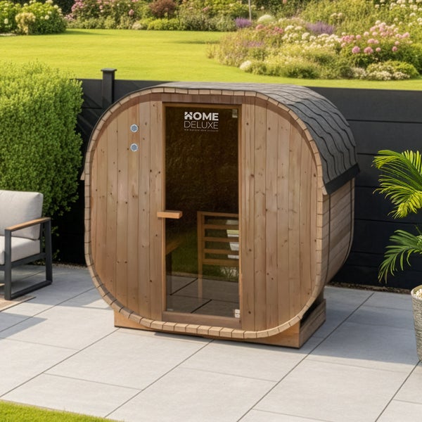 Fasssauna von Home Deluxe mit Glasfront, aus Holz gefertigt, steht auf einer Terrasse in einem gepflegten Garten.
