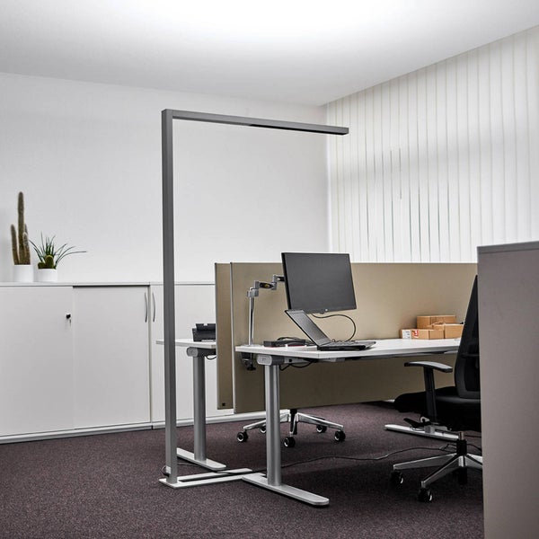 Moderne LED-Stehleuchte für das Büro an einem hellen Arbeitsplatz mit Schreibtisch, Monitor und Bürostuhl.