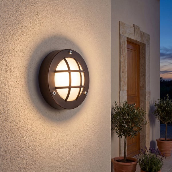 Runde Außenwandleuchte mit Gitterdesign, montiert an einer Hauswand neben einem Eingangsbereich in der Abenddämmerung.