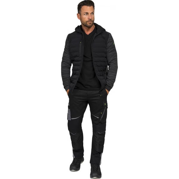 Ein Mann trägt eine schwarze Hybridjacke mit Kapuze, eine schwarze Arbeitshose mit Taschen und dunkle Arbeitsschuhe.