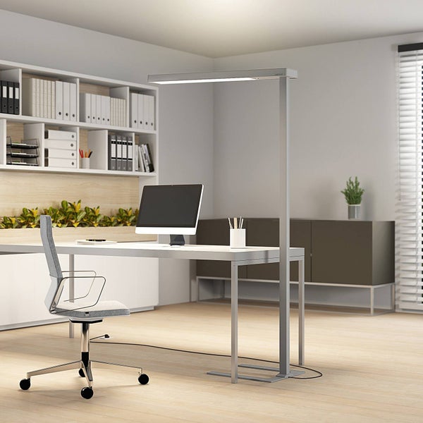 Moderner Büroarbeitsplatz mit weißem Schreibtisch, Monitor, Bürostuhl und einer hohen silbernen LED-Stehleuchte in hellem Ambiente.