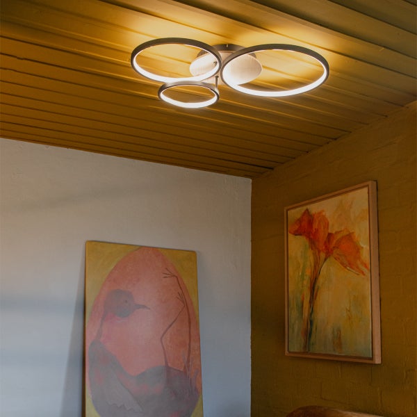Moderne LED Deckenleuchte mit drei ringförmigen Lichtelementen in einem Wohnraum mit Wandbildern und Holzelementen.