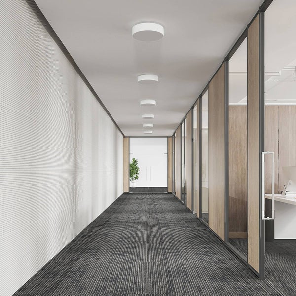 Moderner, heller Bürogang mit dunklem Teppichboden, runden Deckenlampen und Glaswänden mit Holzelementen.