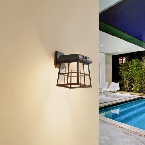 Beleuchtete schwarze Außenwandleuchte im Laternen-Design an einer hellen Fassade neben einem modernen Poolbereich bei Nacht.