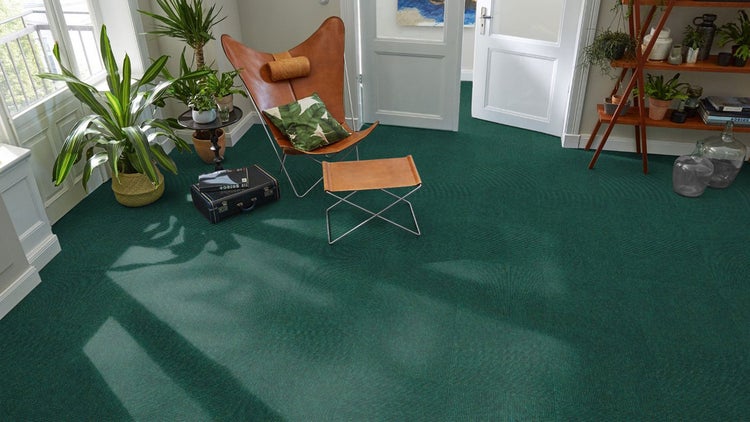 Modernes Wohnzimmer mit dunkelgrünen, texturierten Teppichfliesen, einem braunen Butterfly-Sessel aus Leder und diversen Zimmerpflanzen.