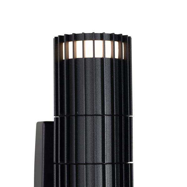 Zylindrische Außenwandleuchte aus schwarzem Metall mit vertikalen Lamellen und warmweißem Lichtaustritt am oberen Rand.