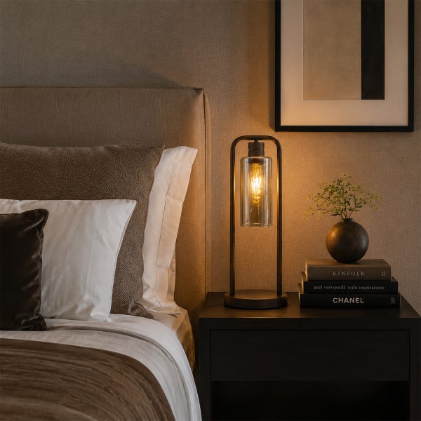 Moderne Tischleuchte mit Metallrahmen und zylindrischem Glasschirm auf einem Nachttisch im Schlafzimmer mit dekorativen Büchern und einer Pflanze.