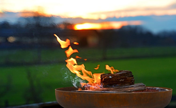 Eine Feuerschale aus Metall mit brennendem Kaminholz im Garten bei Sonnenuntergang.