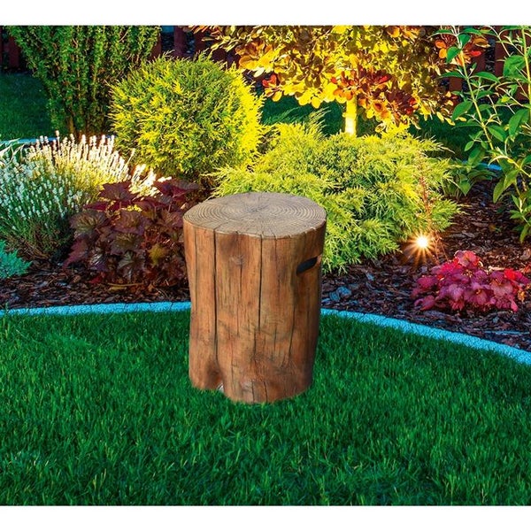 Gartensteckdose in Baumstumpf-Optik auf einer Rasenfläche vor einem beleuchteten Gartenbeet mit Sträuchern.