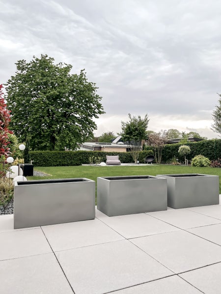 Drei große rechteckige Pflanzgefäße aus Fiberstone auf einer modernen Terrasse in einem gepflegten Garten mit Rasen und Hecken.