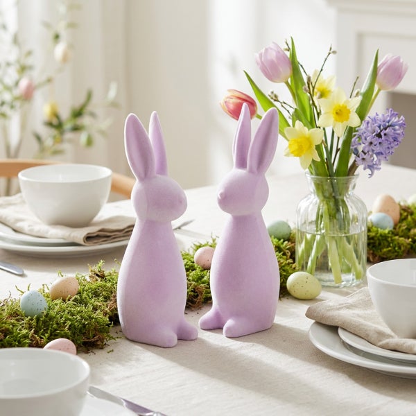 Zwei lilafarbene Osterhasen Figuren aus beflocktem Material auf einem festlich gedeckten Tisch mit Blumen und Ostereiern als Osterdekoration.