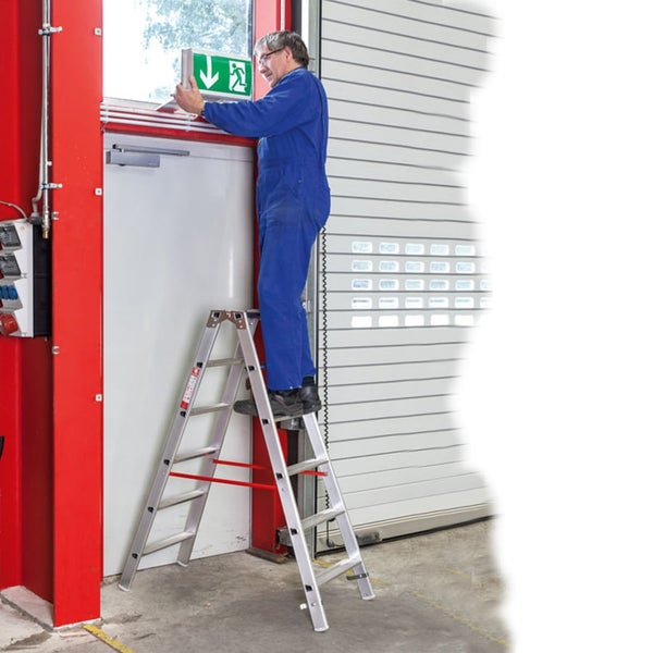 Ein Handwerker steht auf einer Aluminium-Doppelstufenleiter und montiert ein Notausgang-Schild über einer Tür in einer Werkstatt.