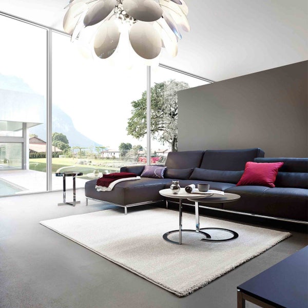 Modernes Wohnzimmer mit dunklem Ledersofa, hellem Teppich und runden Couchtischen vor bodentiefen Fenstern mit Ausblick ins Grüne.