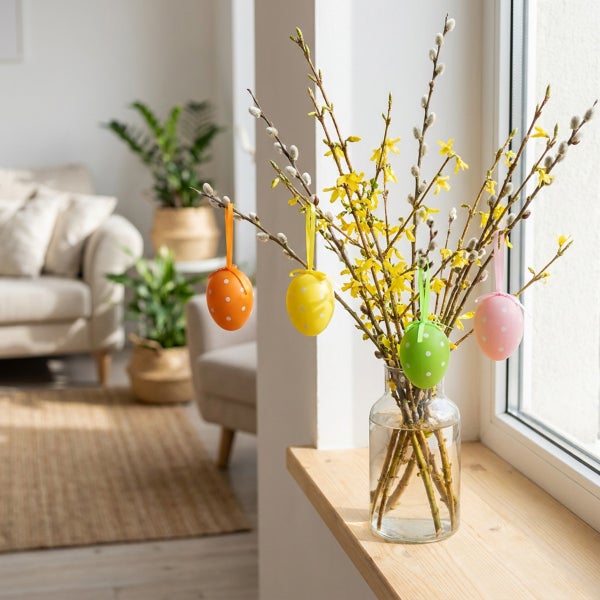 Glasvase mit Forsythienzweigen und hängenden Ostereiern als Dekoration auf einer Fensterbank in einem hellen Wohnraum.