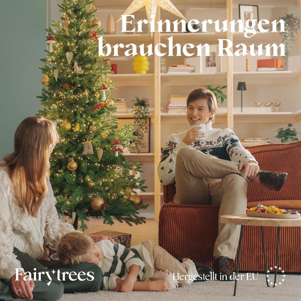 Familie feiert Weihnachten in einem gemuetlichen Wohnzimmer mit geschmuecktem Fairy Trees Weihnachtsbaum und Regalsystem.