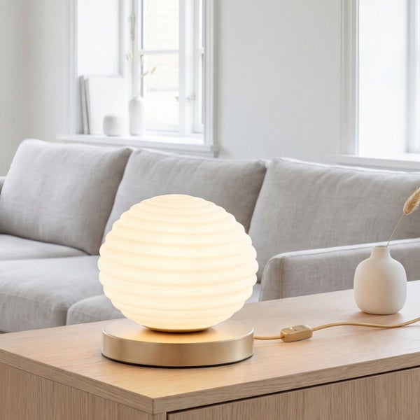 Moderne Tischleuchte mit geriffeltem Schirm und Metallsockel auf einem Holztisch in einem hellen Wohnzimmer mit Sofa.