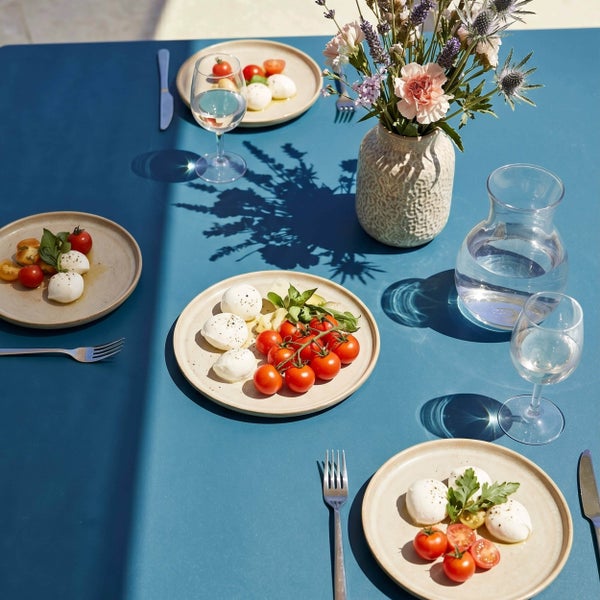 Gedeckter Gartentisch aus HPL in Blau mit Tellern, Besteck, Gläsern, einer Wasserkaraffe und einer Blumenvase im hellen Sonnenlicht.