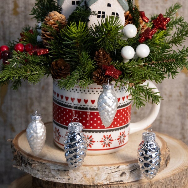 Weihnachtsdekoration mit Tassengefäß, Tannenzweigen, Zapfen und hängenden Christbaumkugeln in Zapfenform auf einer Holzscheibe.