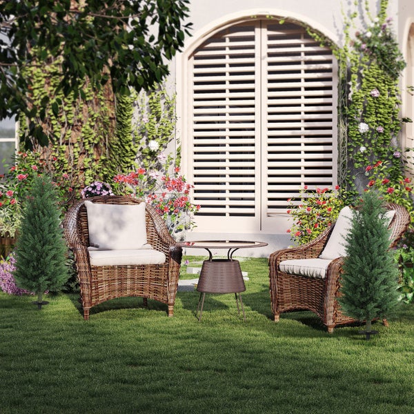 Zwei Rattansessel mit hellen Polstern, ein runder Glastisch und zwei künstliche Zypressen auf einer Rasenfläche vor einer bewachsenen Fassade.