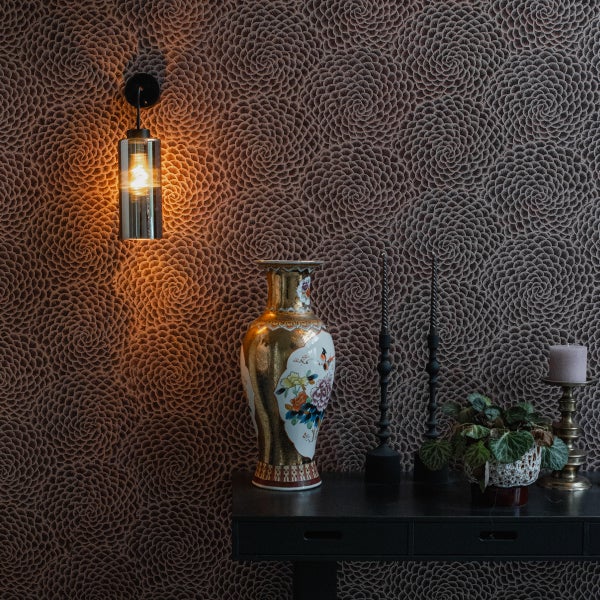 Eine Wand mit gemusterter Tapete in Spiraloptik, eine Wandleuchte aus Glas mit warmem Licht und ein Sideboard mit einer verzierten Blumenvase.