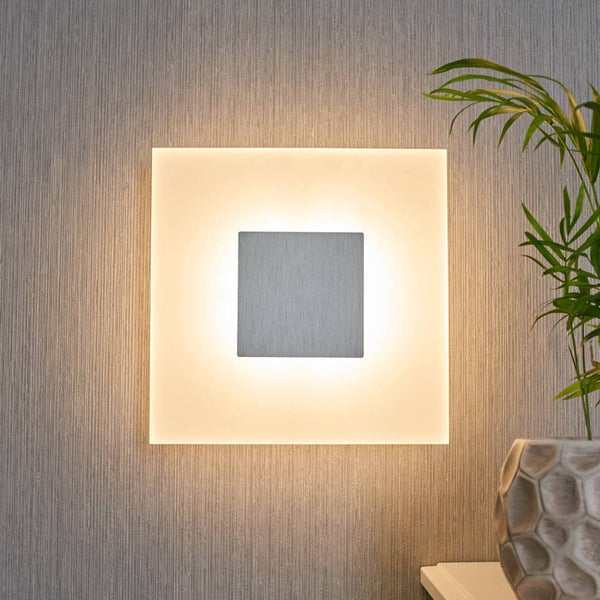 Quadratische LED-Wandleuchte mit Metallplatte und warmem Hintergrundlicht an einer strukturierten Wand neben einer Zimmerpflanze.