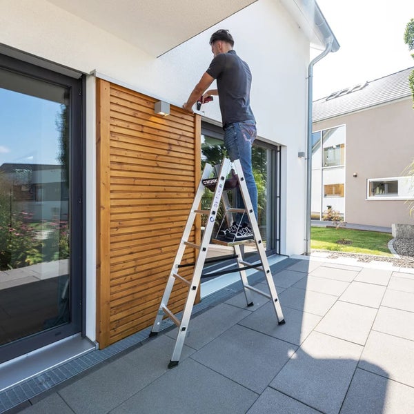 Handwerker arbeitet auf einer Aluminium Stufenstehleiter an einer Holzelement Fassade vor einem modernen Haus.