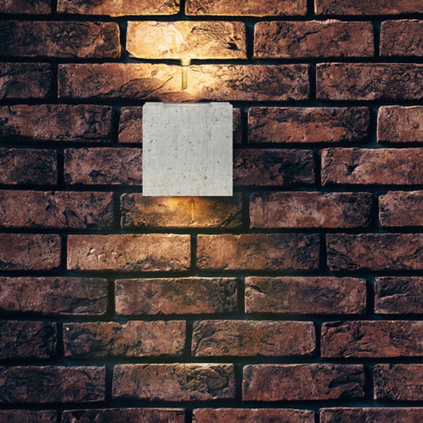 Quadratische Wandleuchte aus Beton mit Lichtaustritt nach oben und unten, montiert an einer rustikalen Ziegelwand.