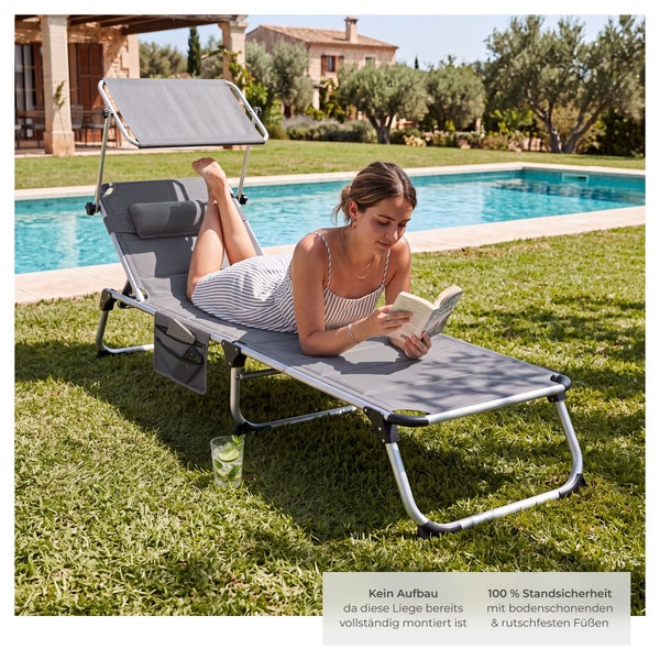 Eine Frau liegt auf einer grauen Gartenliege mit Sonnendach und liest ein Buch auf einer Rasenfläche neben einem Pool.