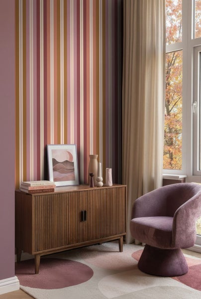 Modern eingerichtetes Zimmer mit mehrfarbig gestreifter Tapete, einem Sideboard aus Holz, einem violetten Sessel und einem gemusterten Teppich.