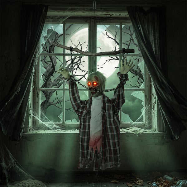 Hängende Zombie Halloween Dekofigur mit leuchtenden Augen in einem Fenster mit gebrochenen Scheiben und Ketten.