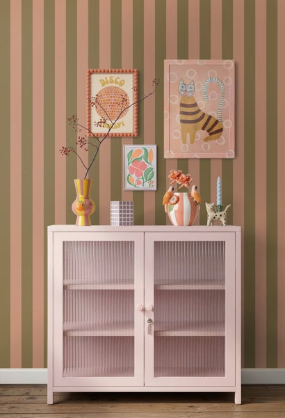 Gestreifte Tapete in Olivgrün und Altrosa hinter einem rosa Metallschrank mit Riffelglas und bunter Dekoration.