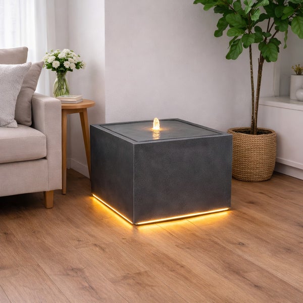 Würfelförmiger Zimmerbrunnen in Grau mit LED-Beleuchtung am Sockel und beleuchtetem Wasseraustritt in einem hellen Wohnzimmer.