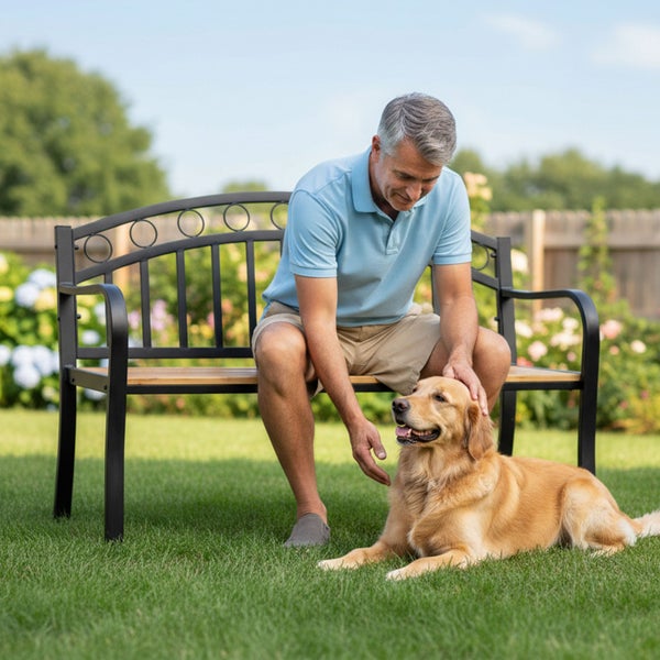 Gartenbank aus schwarzem Metall und Holz in einem hellen Garten, auf der ein Mann sitzt und einen Golden Retriever auf dem Rasen streichelt.