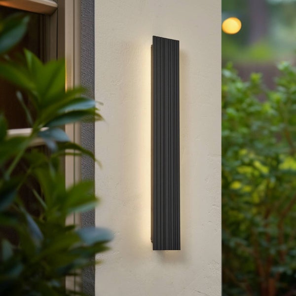 Moderne vertikale Außenwandleuchte mit schwarzer, gerippter Oberfläche und warmweißem seitlichem Lichteffekt, montiert an einer Außenwand.