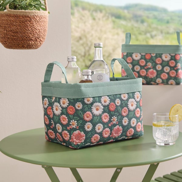 Rechteckiger Aufbewahrungskorb aus Stoff mit floralem Gänseblümchen-Muster auf einem grünen Außentisch mit Glasflaschen.