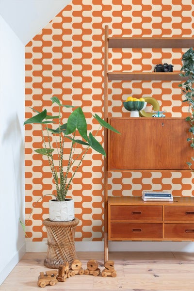 Hell eingerichtetes Zimmer mit retro-gemusterter Tapete, einem Holzregal, einer Zimmerpflanze auf einem Korbhocker und Spielzeug auf dem Fußboden.