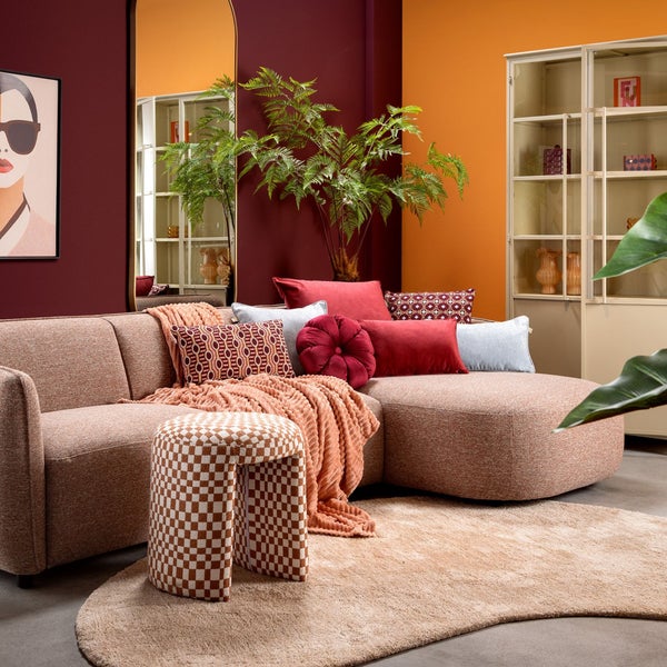 Modernes Wohnzimmer mit stoffbezogenem Ecksofa, gemusterten Dekokissen, strukturiertem Hocker, weichem Teppich und großer Zimmerpflanze.