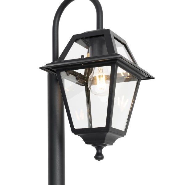 Schwarze Außenleuchte in Laternenform mit Klarglas und leuchtendem Leuchtmittel an einem gebogenen Pfosten.
