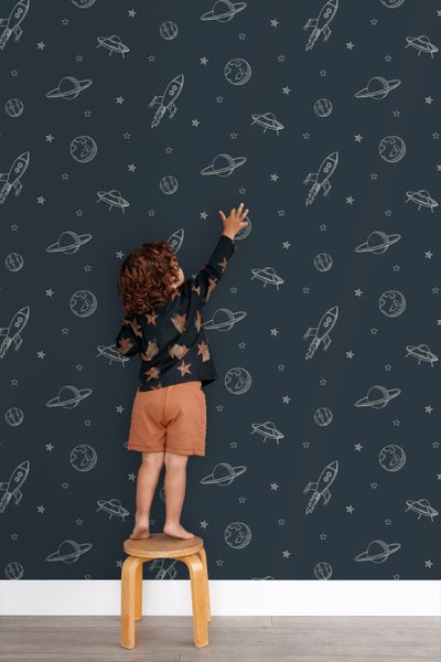 Ein Kind steht auf einem Hocker vor einer Wandtapete mit Weltraummotiven.
