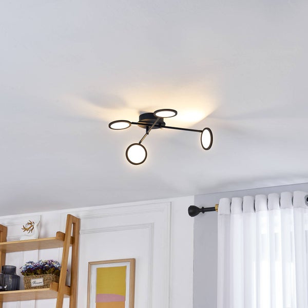 Moderne Deckenlampe aus schwarzem Metall mit vier LED-Ringen, in einem hellen Raum mit Holzmöbeln und weißen Vorhängen.