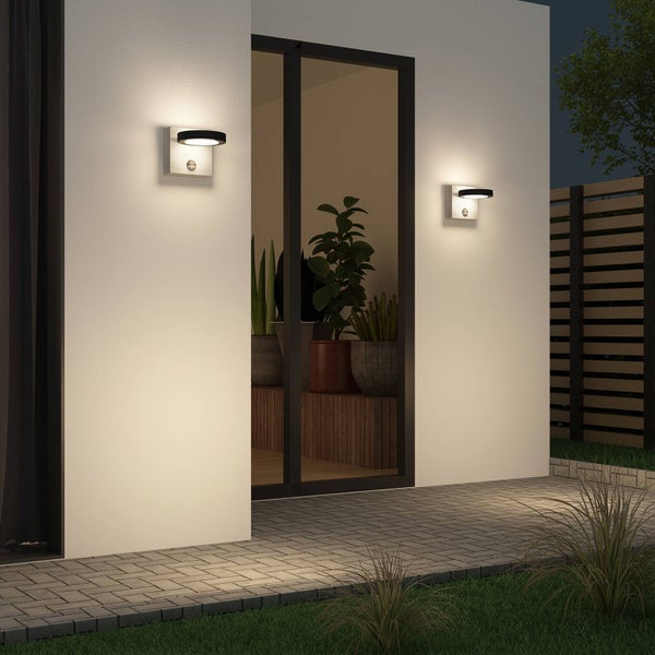 Zwei moderne LED-Außenwandleuchten mit Bewegungsmelder beleuchten eine Wand über einer gepflasterten Terrasse bei Nacht.